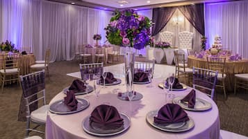 A banquet hall with round tables set for a formal event, featuring white tablecloths, purple napkins, and floral centerpieces.