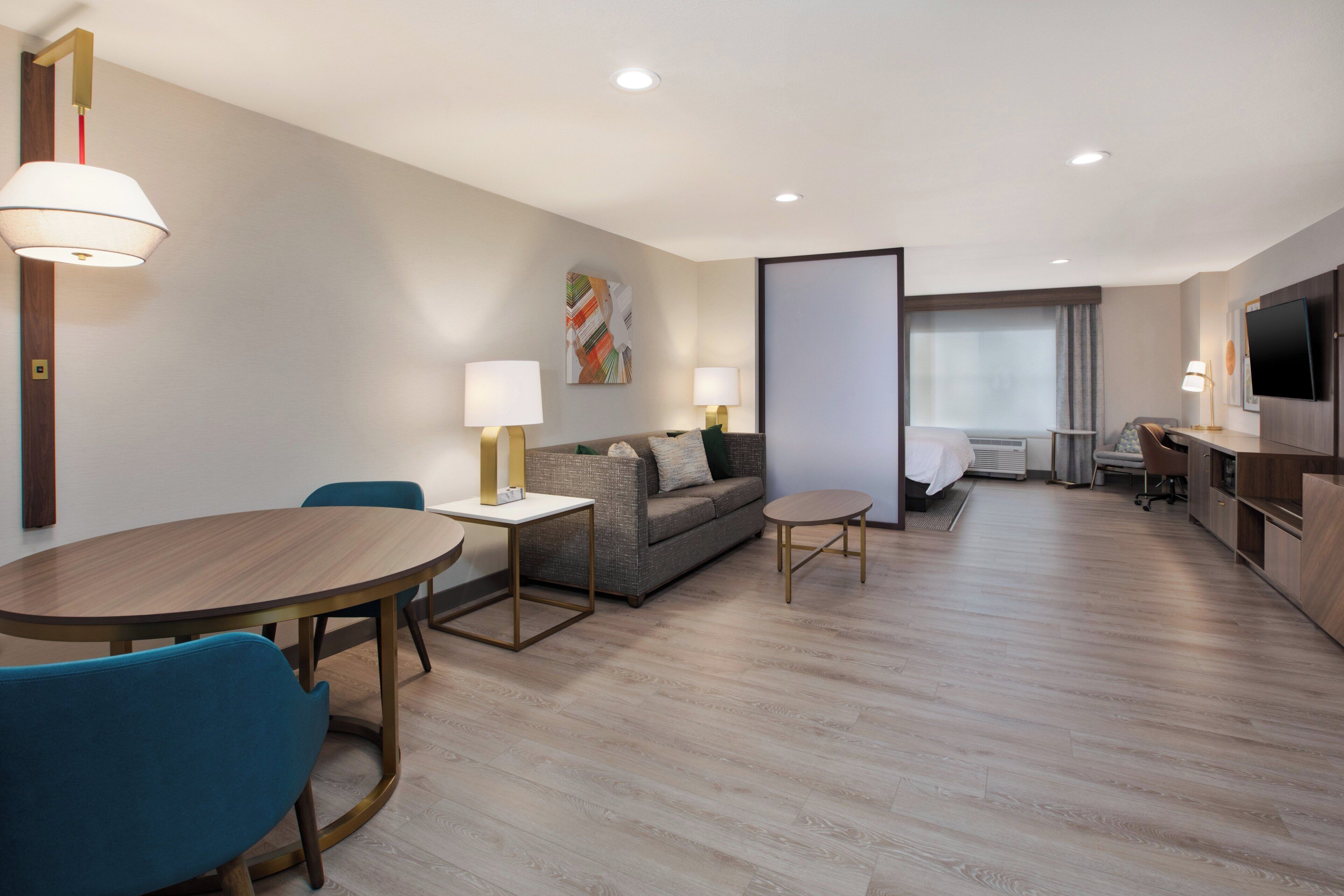 A modern hotel room with a dining area, a sofa, a TV, and a large window.