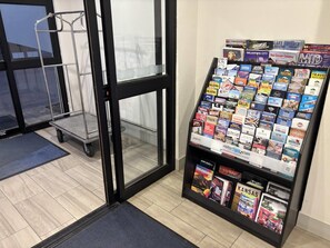 Un área de recepción con puerta de vidrio, un carrito metálico y una exhibición de folletos de viaje.