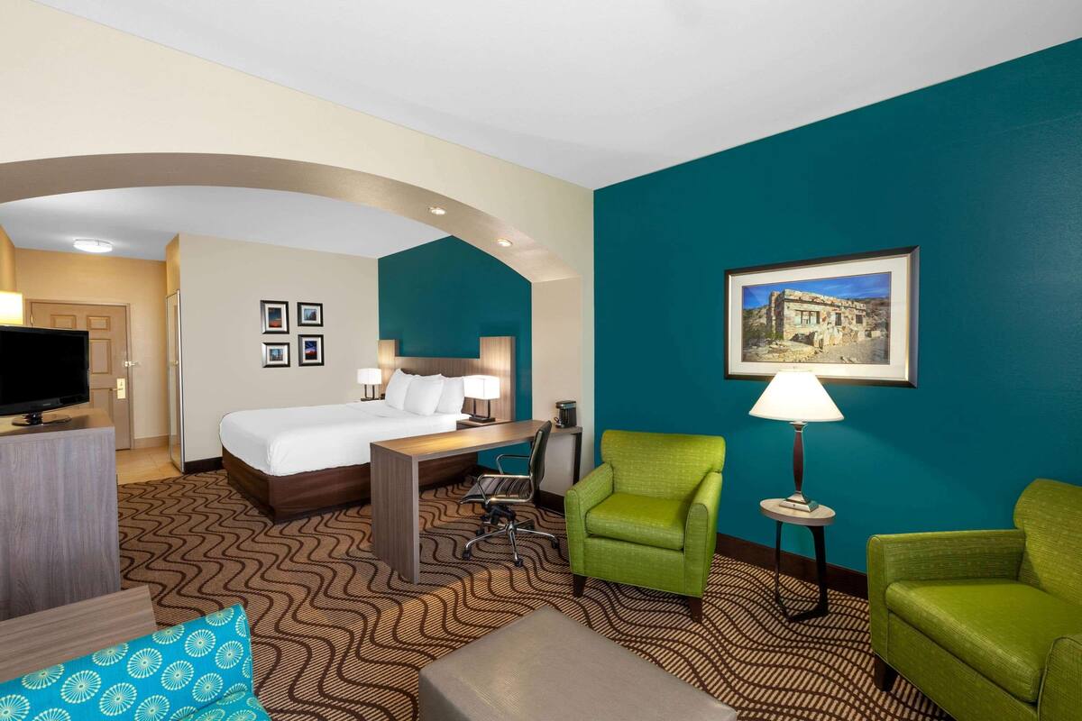 Ein Hotelzimmer mit Bett, Schreibtisch, grünen Sesseln, einem Fernseher und einem gerahmten Bild an der Wand.