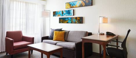 A hotel room with a sofa, armchair, desk, and lamp. There are three paintings on the wall.