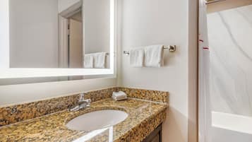 Une salle de bain avec un lavabo doté d’un plan de travail en granit, un miroir, un porte-serviettes et un espace douche.
