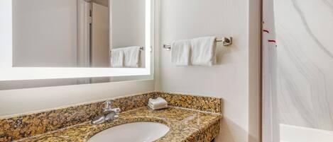 A bathroom with a granite countertop sink, a mirror, a towel rack, and a shower area.