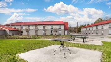 A building with a red roof and multiple windows, a concrete patio with a table and benches, and a grassy area in front.