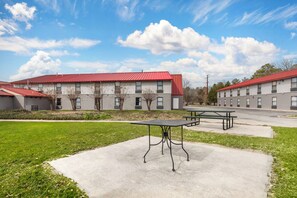 Ein Gebäude mit rotem Dach und zahlreichen Fenstern, eine Betonterrasse mit Tisch und Bänken sowie eine Rasenfläche im Vordergrund.