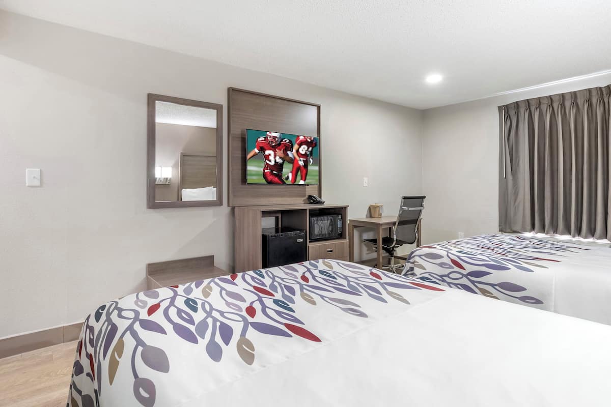 Habitación de hotel con dos camas, un televisor montado en la pared, un escritorio con una silla y una ventana con cortinas.