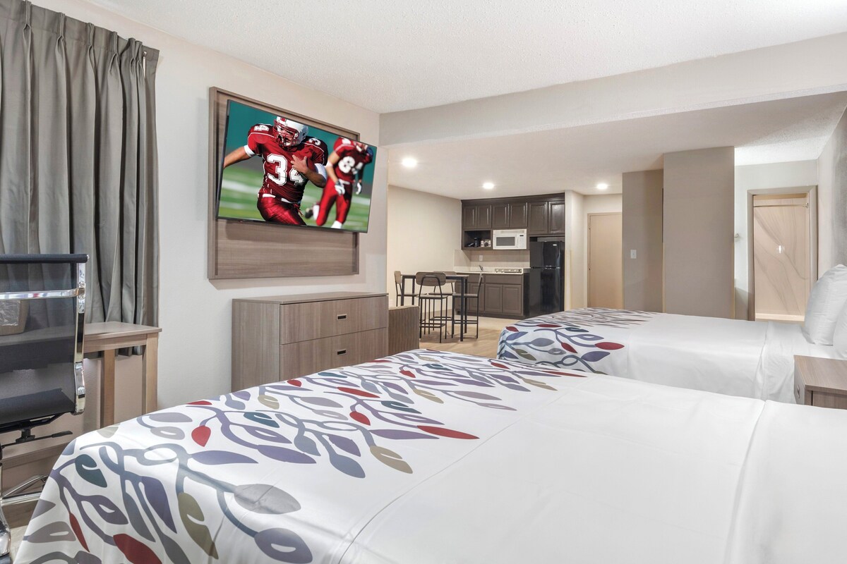 Ein Hotelzimmer mit zwei Betten, einem Fernseher, einem Schreibtisch, einem Sessel und einer Küchenzeile.