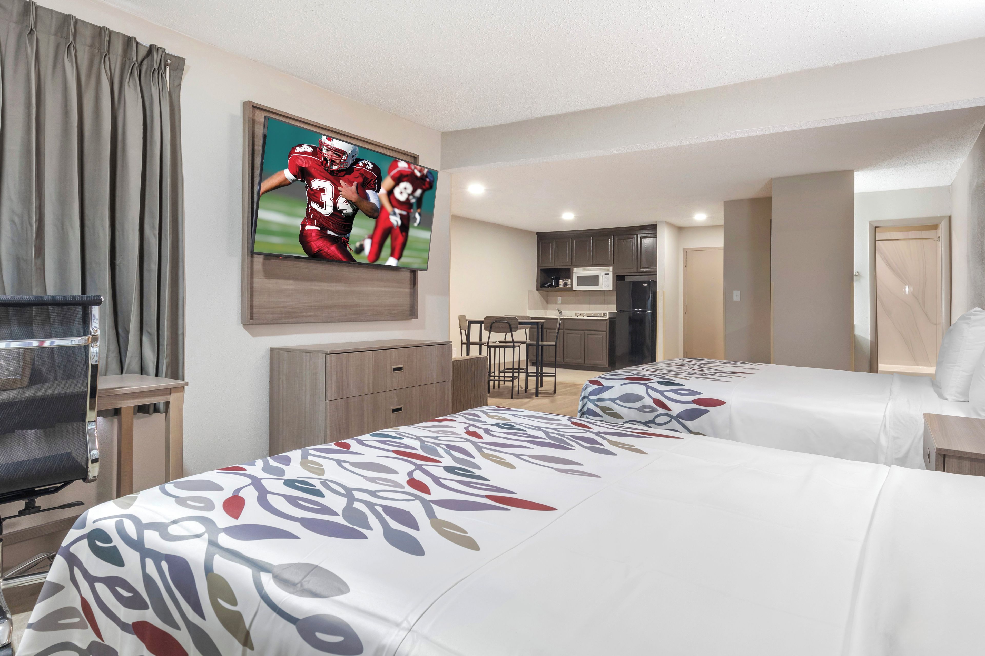 A hotel room with two beds, a TV, a desk, a chair, a kitchenette, and a door.