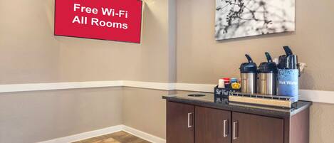 Um saguão de hotel com sinalização indicando Wi-Fi gratuito em todos os quartos, um espaço com café e chaleiras, e uma obra de arte pendurada na parede.