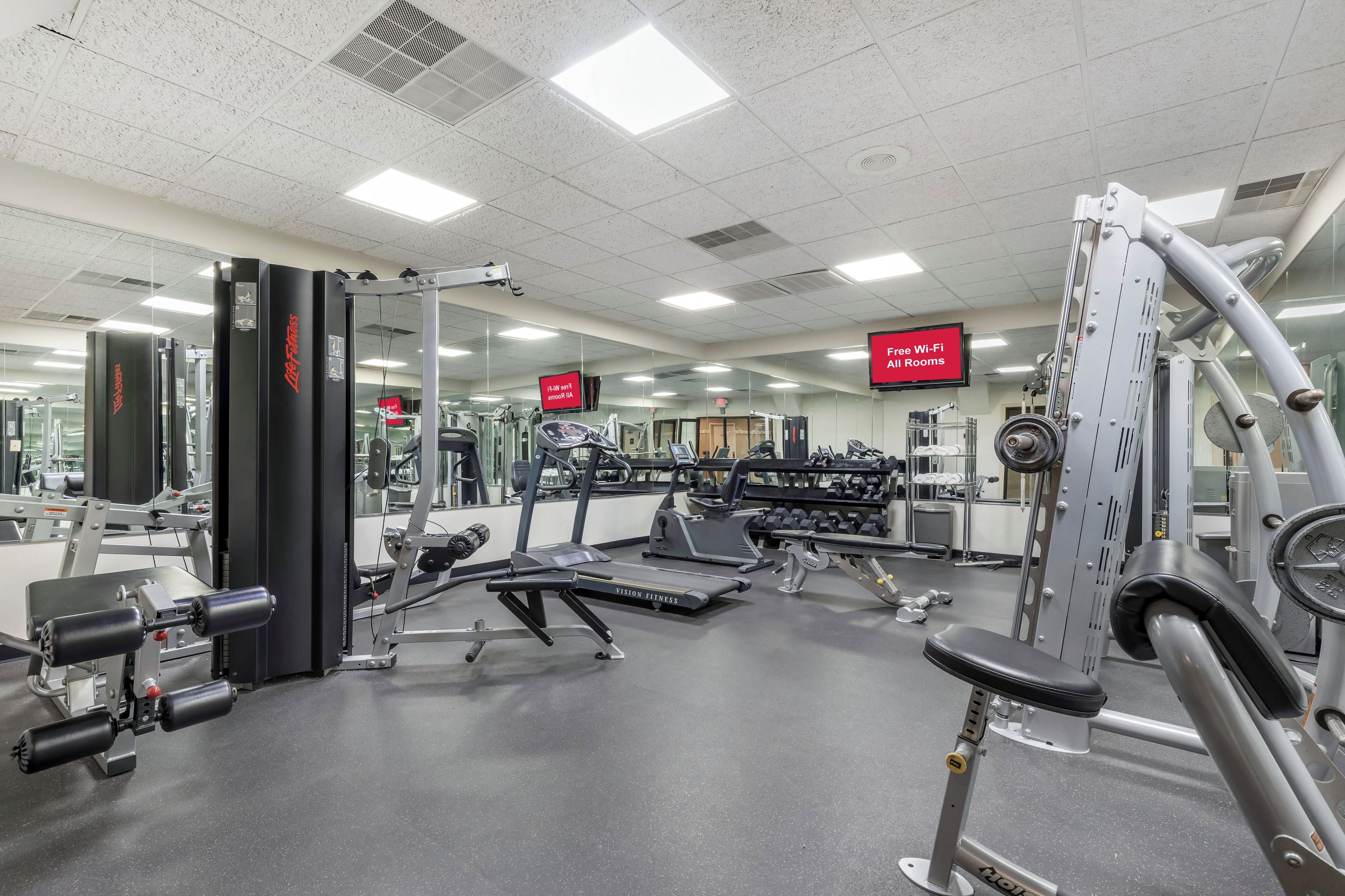 A gym with various exercise equipment, a mirrored wall, and a sign indicating free Wi-Fi in all rooms.