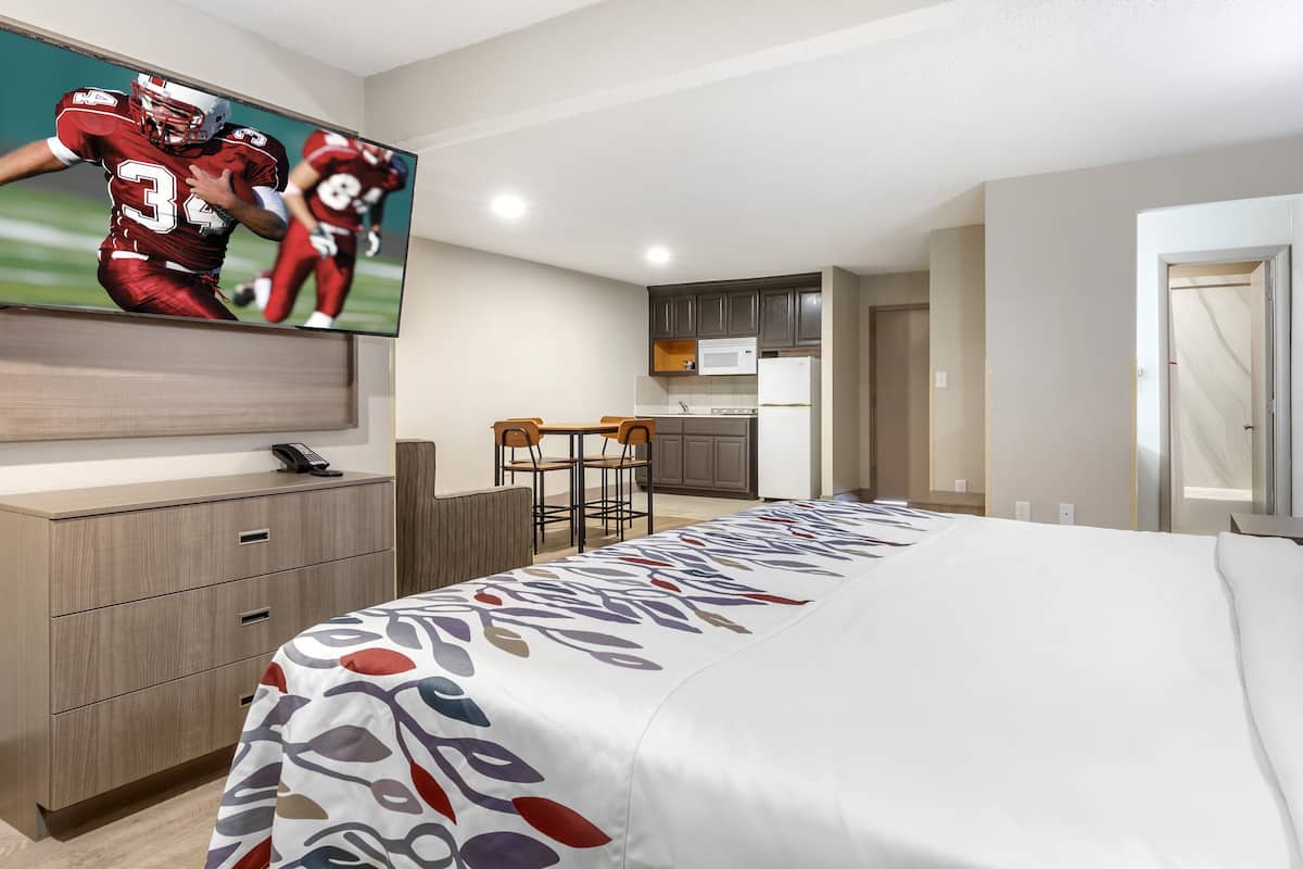 Una habitación de hotel con cama, escritorio, televisor, kitchenette y zona de comedor.