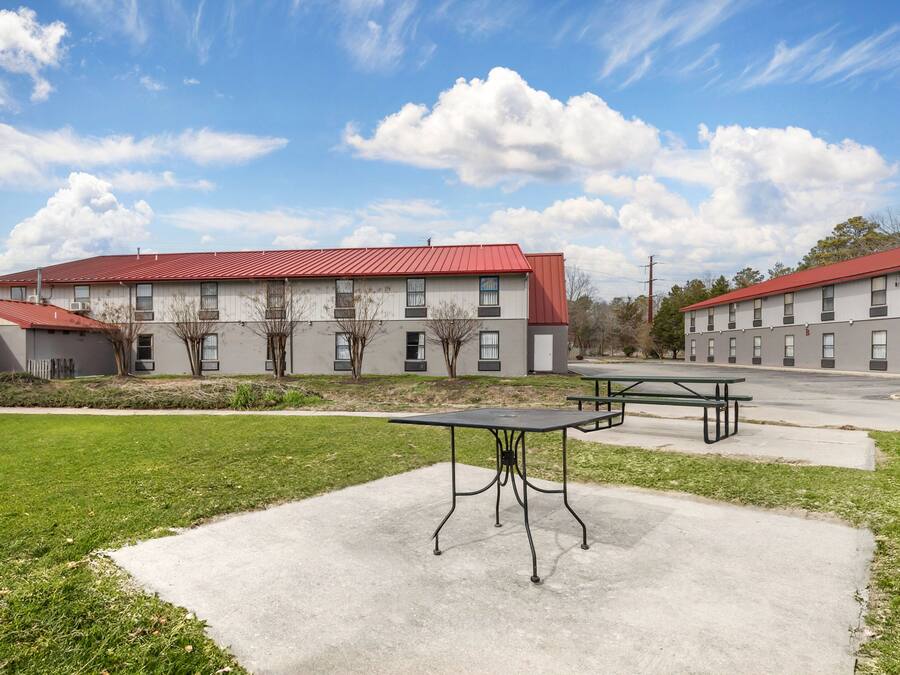 Un edificio con techo rojo y múltiples ventanas, un patio de concreto con una mesa y bancos, y un área verde al frente.