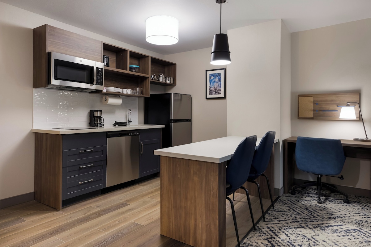 A modern kitchen with a microwave, refrigerator, and wooden cabinets. There is a small island with bar stools and a desk with a chair.