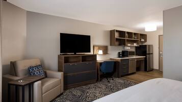 Habitación de hotel compacta con cama, sofá, escritorio, silla, televisor, kitchenette y un mueble pequeño.