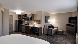 A compact hotel room with a kitchenette, a bed, a sofa, a small table, and a TV.