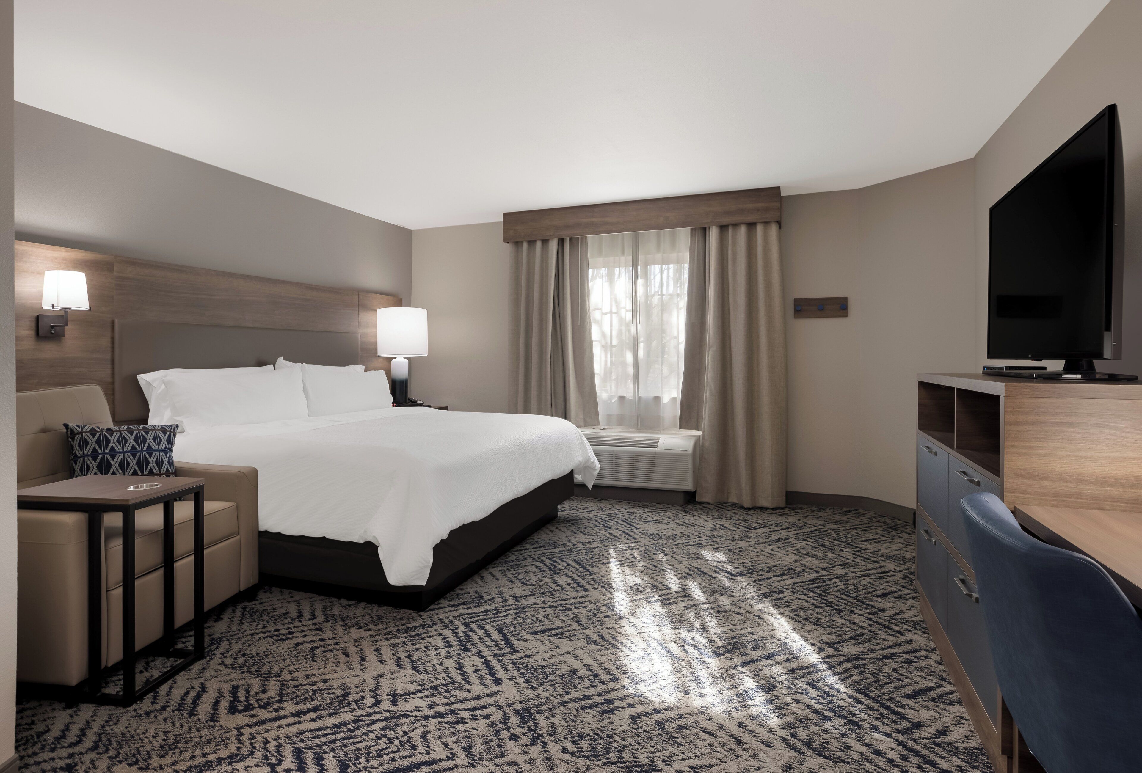 A hotel room with a bed, bedside tables, a desk, a chair, a television, and a window with curtains.