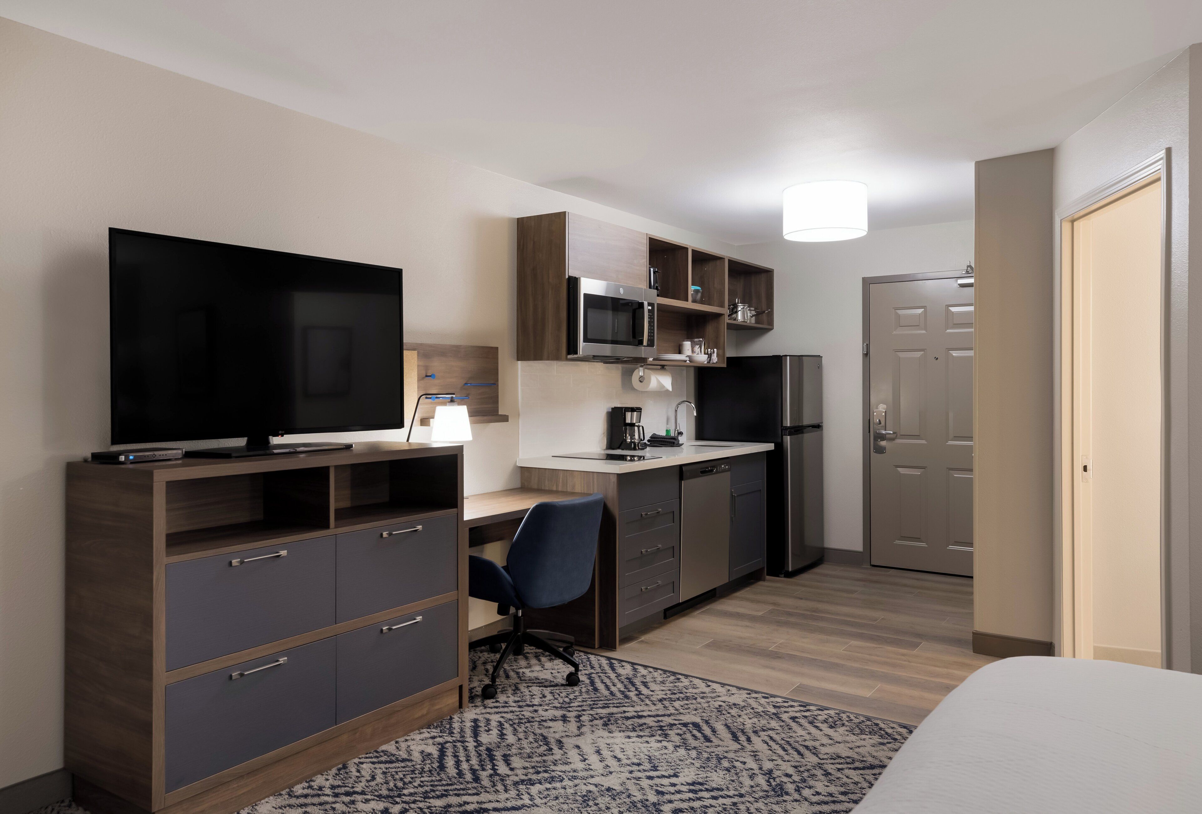 A compact hotel room with a kitchenette, a television, a desk, and a chair.