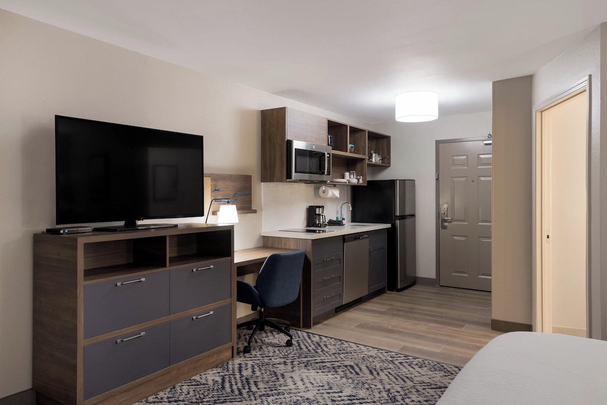 A compact hotel room with a kitchenette, a flat-screen TV, a desk, and a chair.