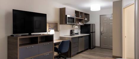 A compact hotel room with a kitchenette, a television, a desk, and a chair.