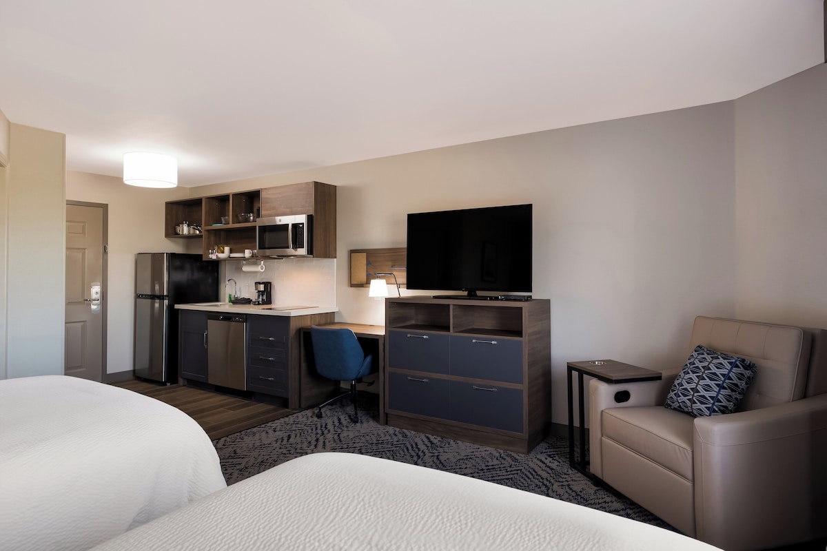 A hotel room with a bed, a desk with a chair, a kitchenette, a TV, and a small seating area.