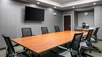 Un salón de conferencias con una larga mesa de madera, seis sillas de malla negra, un televisor de pantalla plana en la pared, un reloj y una cafetera.