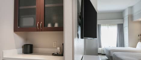 A hotel room with a kitchenette, a TV mounted on the wall, two beds, and a window with curtains.