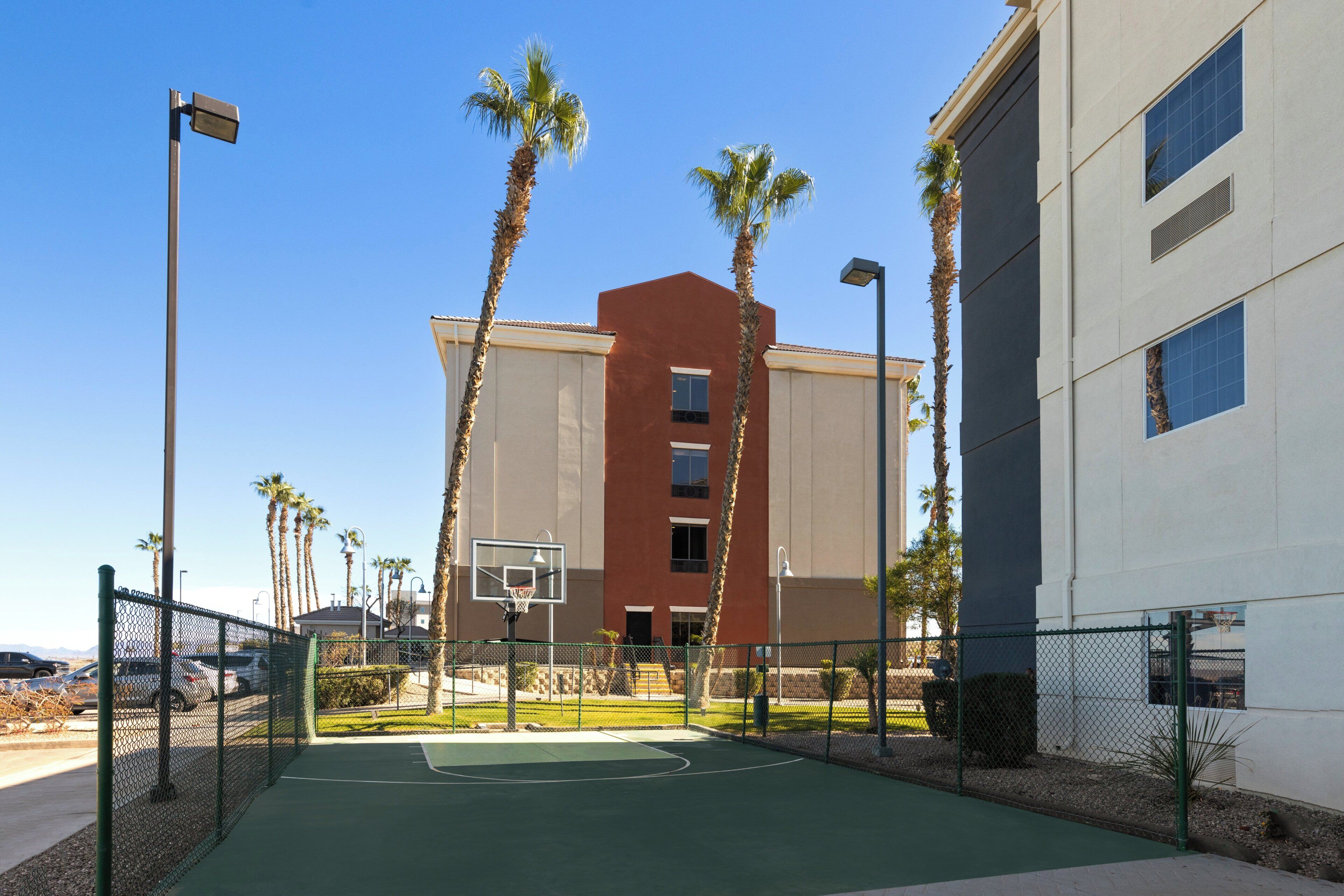 An outdoor basketball court with a chain-link fence, palm trees, and a building with a red facade in the background.