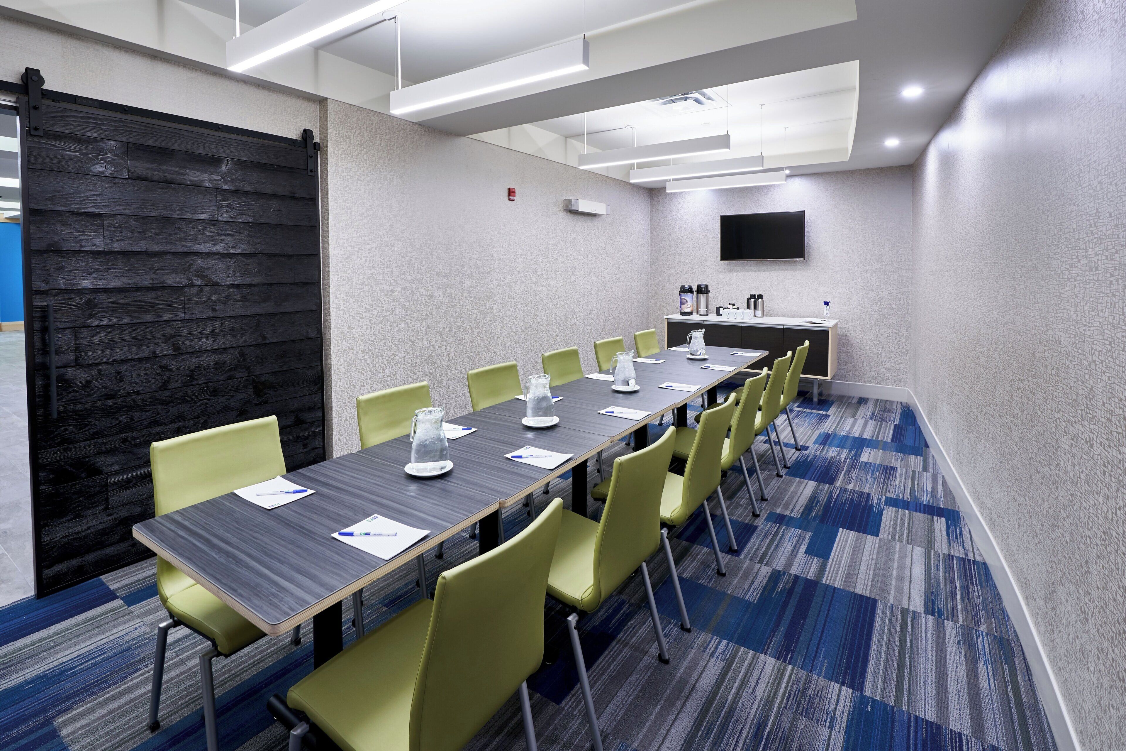 A conference room with a long table, green chairs, a black wall, and a mounted TV.
