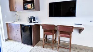 Cocina compacta con lavavajillas, armarios de madera, microondas y televisor de pantalla plana. Hay dos sillas de madera y una mesa pequeña blanca.