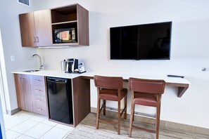 Una cucina compatta con lavastoviglie, mobili in legno, forno a microonde e TV a schermo piatto. Si notano due sedie in legno e un piccolo tavolo bianco.