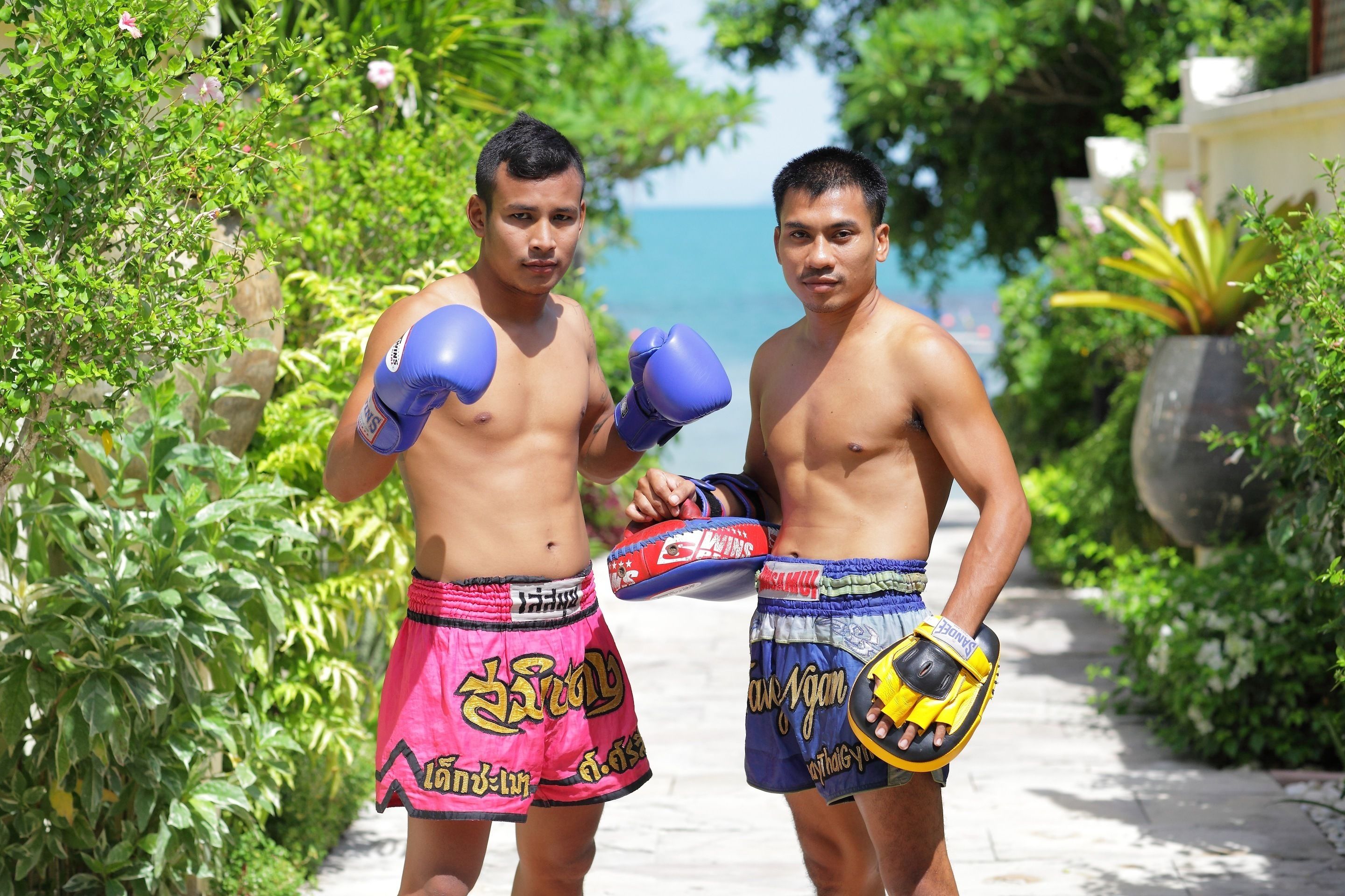 Dos personas con shorts deportivos y guantes de boxeo de pie en un sendero, con vegetación y vista al mar.