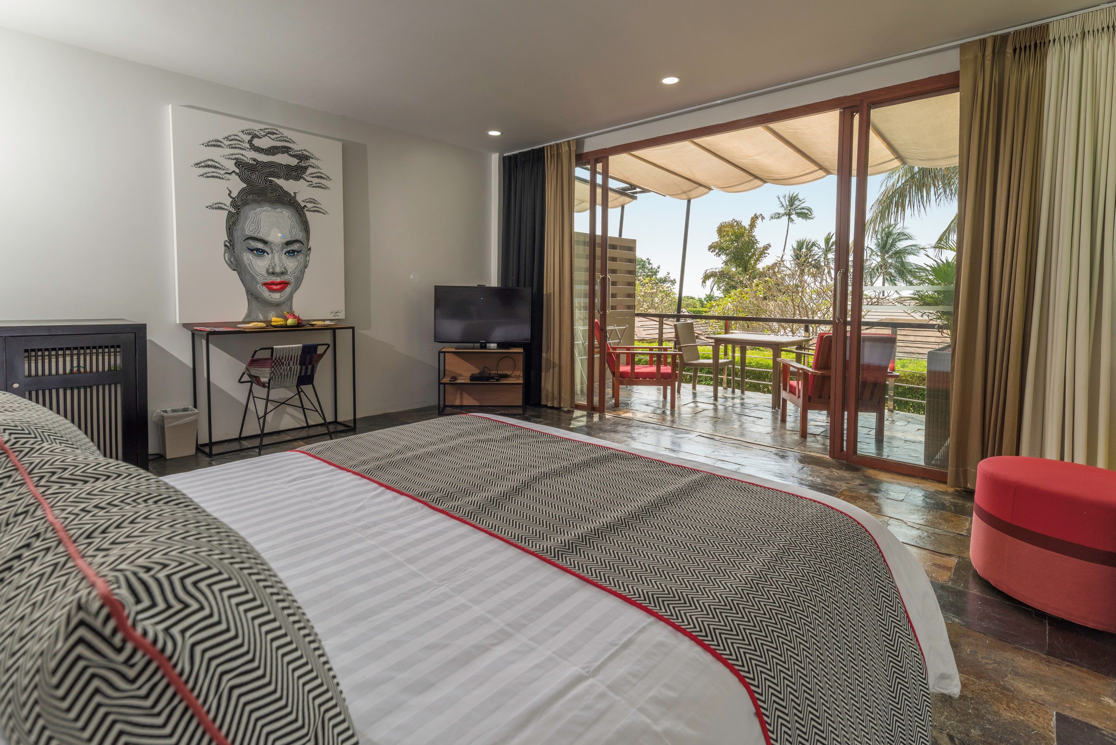A hotel room with a large bed, a TV, a desk with a chair, and a balcony with outdoor seating.