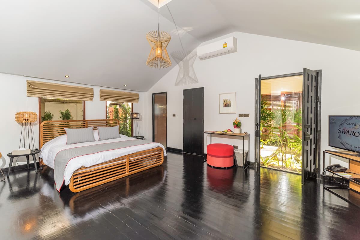 A modern bedroom with a wooden bed, a red ottoman, a desk with a computer, and a large window with a view of greenery.