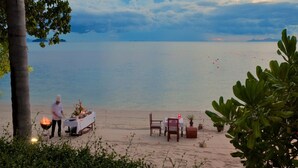 A beachside dining setup with a table and chairs, a chef preparing food, and a view of the sea. - The Sunset Beach Resort & Spa Taling Ngam (Koh Samui)