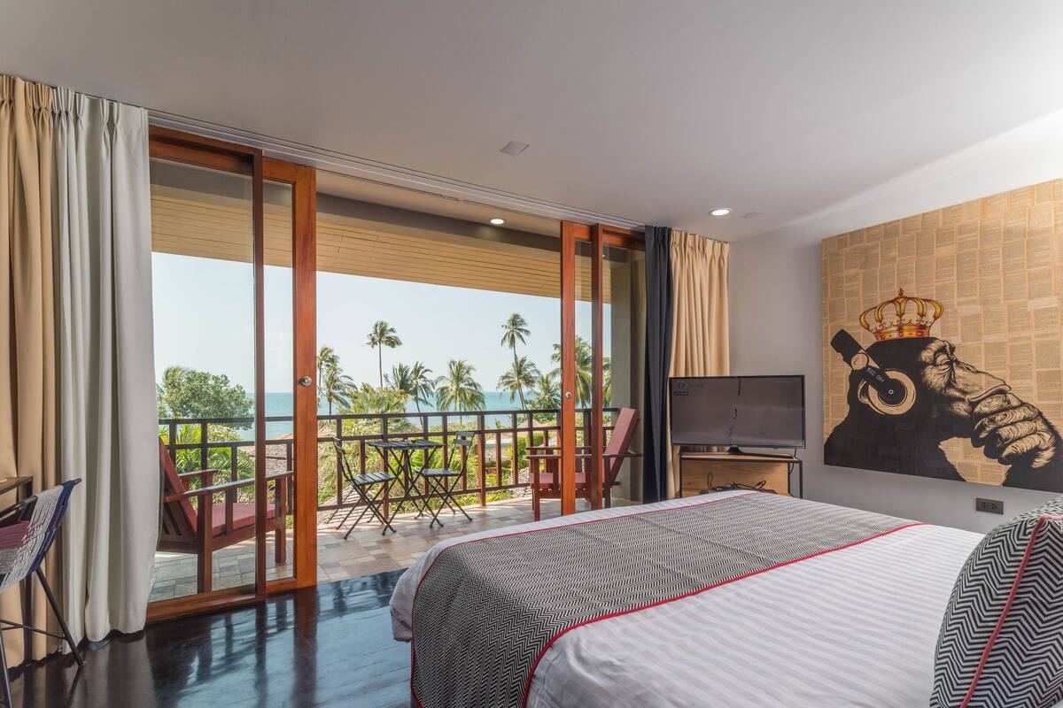 A hotel room with a large bed, a TV, a balcony with a view of the sea, and a painting on the wall.