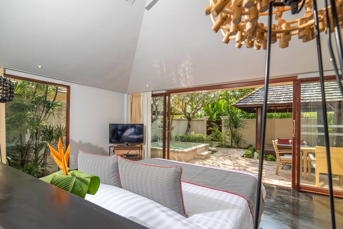 A modern hotel room with a large bed, a TV, and a view of the outdoor garden and patio area.