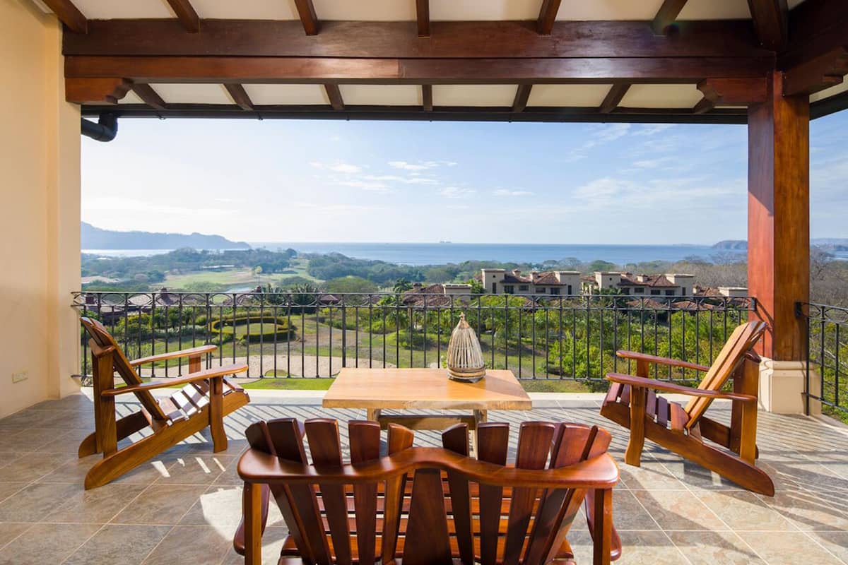 A covered outdoor seating area with a table and chairs, overlooking a scenic landscape with buildings and greenery.