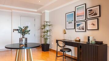 Een moderne hotelkamer met een houten vloer, een ronde tafel met een bloempot, een zwarte stoel, een bureau met een lamp en lijstjes met foto's aan de muur.