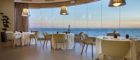Un comedor con mesas redondas, sillas e iluminación colgante, con vistas a una masa de agua.