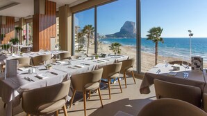 Un restaurante con mesas dispuestas para comer, grandes ventanales que ofrecen vista a la playa y al océano.