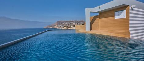 A modern infinity pool with a clear view of the sea and a coastal town in the background.