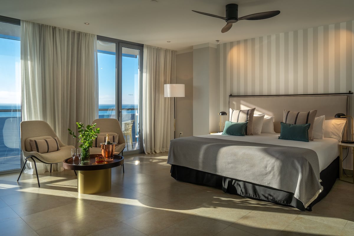 A modern hotel room with a large bed, a sitting area with a coffee table, and a view of the ocean through floor-to-ceiling windows.