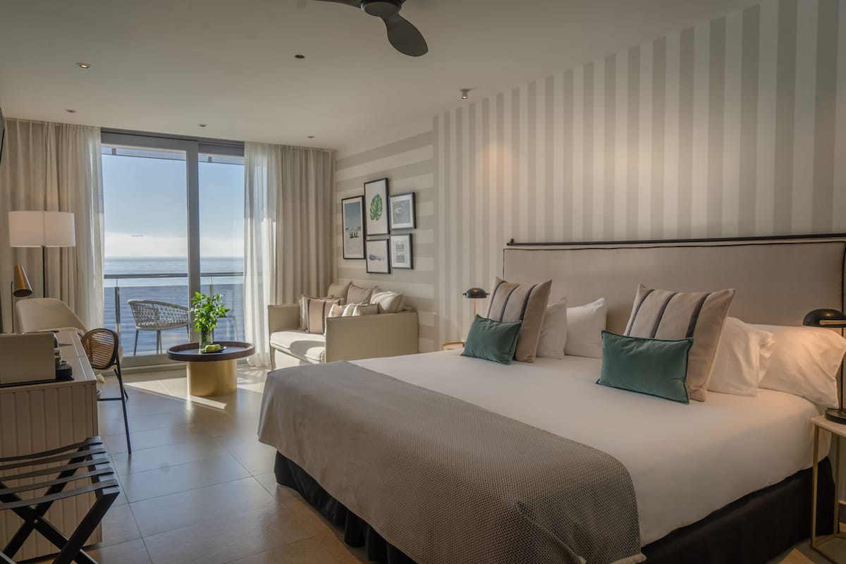 A modern hotel room with a large bed, a seating area with a view of the sea, and a balcony.
