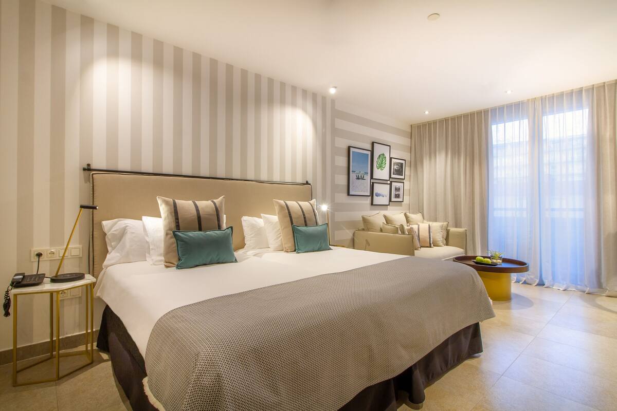 A hotel room with a large bed, a bedside table with a lamp, a small table with a plant, and a window with curtains.