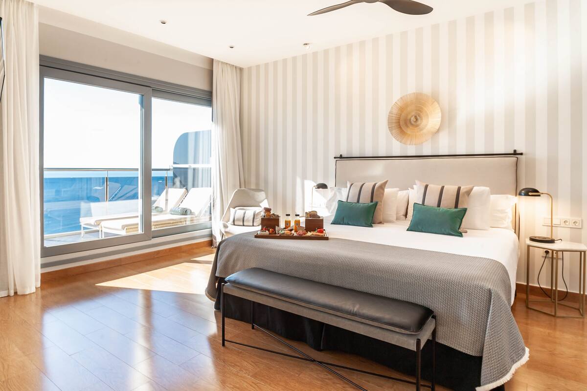 A modern hotel room with a large bed, a bench, a side table, and a view of the ocean.