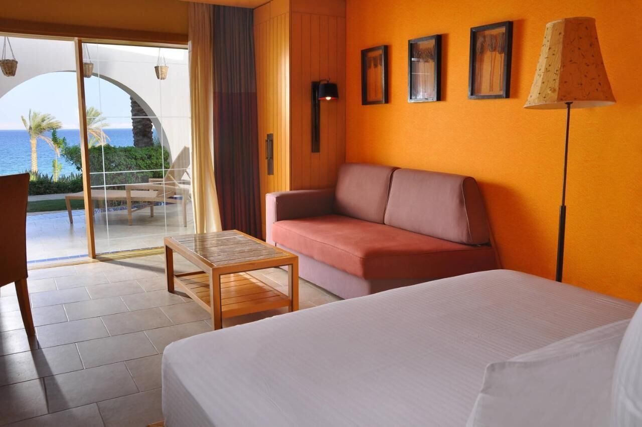 A hotel room with a bed, a sofa, a small table, and a view of the ocean through a glass door.