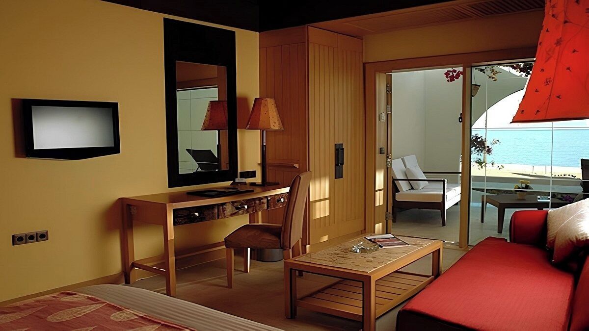 A hotel room with a bed, a desk, a chair, a small table, a TV, and a balcony with a view of the sea.