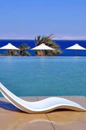 Zona de piscina con una silla de descanso, palmeras y sombrillas.