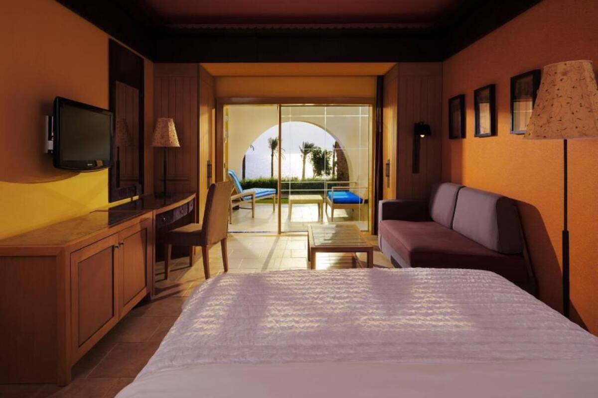 Ett hotellrum med en säng, en TV, ett skrivbord, en stol, en soffa, en lampa och utsikt över poolen och palmerna.