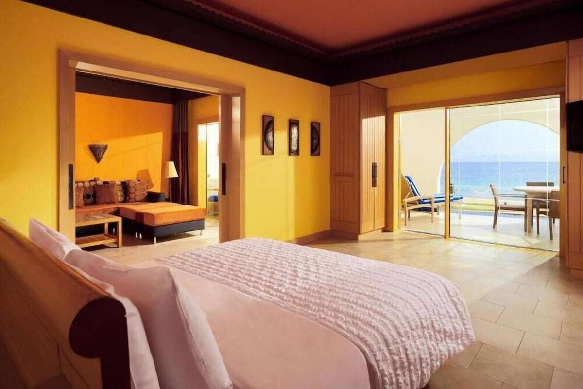 Ett hotellrum med en säng, en sittgrupp, utsikt över havet och en balkong med stolar.
